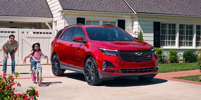 2024 Red Hot Chevy Equinox parked in front of a contemporary white house in Ballinger, Tx with a parent watching their kid ride a bike in the driveway.